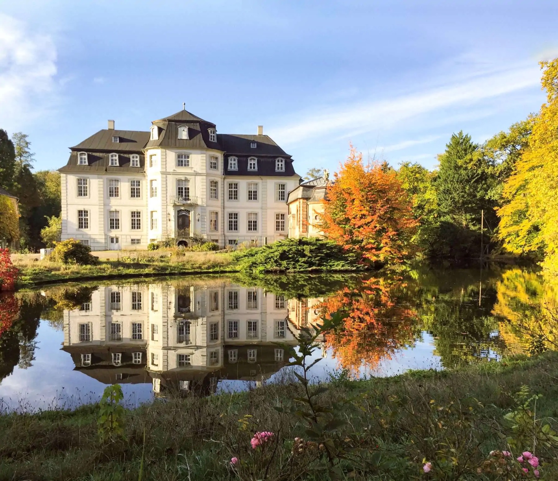 Schloß Türnich spiegelt sich im Weiher, Bäume in Herbstfärbung.